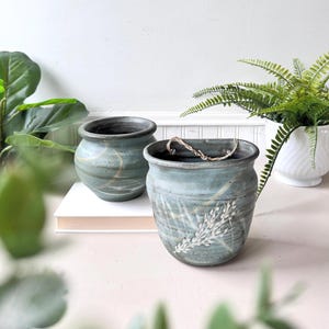 May include: Two handcrafted ceramic planters in shades of blue-green. One is a round pot with gold accents, and the other is a hanging planter with a white floral design. Both have dark rims and are suitable for indoor plants.