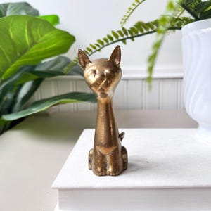 May include: A vintage brass cat figurine. The cat is sitting upright with its ears perked up. The brass has a warm, golden hue. The cat is sitting on a stack of white books. Decorative home decor.
