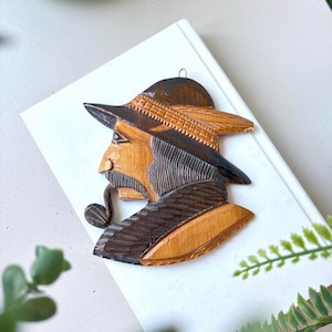 May include: Wooden wall art depicting a man in profile, wearing a hat and smoking a pipe. The carving features intricate details and a mix of light and dark wood tones. The artwork is likely a decorative piece for a home.