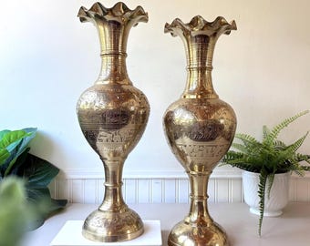 May include: Two tall, polished brass vases with flared, ruffled rims and slender necks. The bulbous bodies feature intricate, etched floral and leaf patterns. The vases stand on circular, pedestal-style bases, showcasing a metallic gold finish.