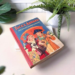 Puede incluir: Libro antiguo "Tuck's Annual" con lomo rojo e ilustración de portada de dos niños bajo un paraguas, uno con un instrumento musical. El texto de la portada dice "Tuck's Annual with Realistic Surprise Panoramas". El libro está ligeramente desgastado.