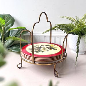 May include: A set of six coasters with a floral design, each with a red border and a cream-colored center. They are held in a gold-colored metal stand with a decorative handle. The stand has a circular base and three curved legs.