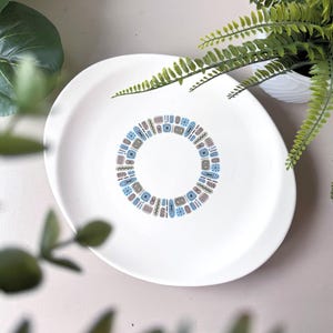 May include: White ceramic plate featuring a decorative ring of blue, brown, and grey geometric shapes. The plate has a wide rim and a central design. The plate is round and appears to be new.
