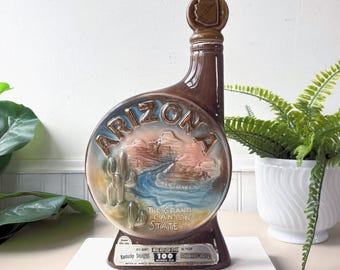 May include: A brown ceramic decanter with a round Arizona-themed design. The design features a desert landscape with the words "ARIZONA" and "THE GRAND CANYON STATE". The decanter has a brown stopper with an Arizona outline.