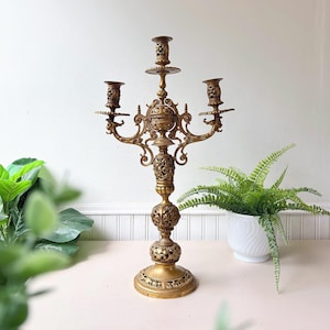 Puede incluir: Un candelabro de metal dorado con tres portavelas, un tallo central ornamentado con detalles de filigrana calada y una base circular ancha. La estructura presenta brazos en voluta y motivos florales decorativos.