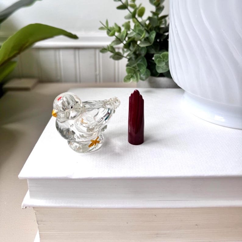 May include: A clear glass bird figurine with a yellow beak and feet, and a red dot on its head. Beside it is a tall, dark red, decorative object. The objects are on a white surface.