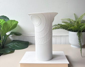 May include: A matte white cylindrical vase featuring a flared top rim and an intricate, raised dot pattern that forms a fan-like design on the upper portion of the exterior. The vase is positioned upright on a rectangular book with a light cover.