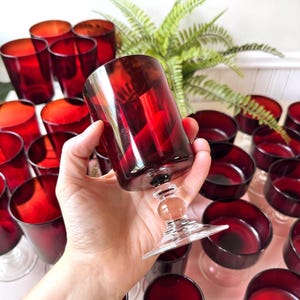 May include: A collection of deep red glass goblets featuring a smooth, cylindrical bowl and a clear, multi-tiered pedestal base. The glassware is uniform in colour and design, with a translucent finish that catches the light.