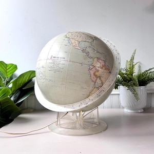 Puede incluir: Un globo terráqueo Time-Life vintage con un mapa de color claro, montado sobre un soporte de plástico transparente. El globo presenta un anillo meridiano blanco y un cable de alimentación. El texto "TIME-LIFE GLOBE" es visible.