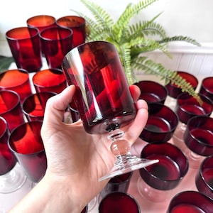 Può includere: Una collezione di calici da vino rosso rubino. I calici hanno una coppa cilindrica, uno stelo decorativo e una base larga. I calici sono disposti su una superficie rosa chiaro, con una felce sullo sfondo.