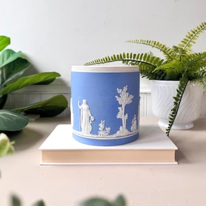 May include: A blue ceramic cylinder with white relief detailing. The design features classical figures and trees. The cylinder is sitting on a white book. A decorative piece for home decor.
