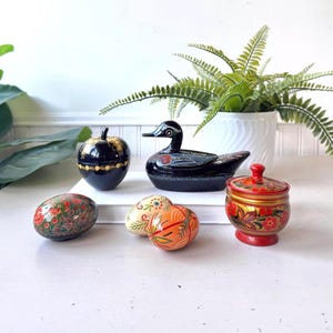 May include: A collection of lacquered objects including a black apple-shaped box, a black duck-shaped box, two small egg-shaped containers with floral patterns, and a red lidded jar with gold and red floral designs on a white surface.