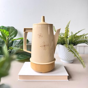 Może przedstawiać: Zabytkowy beżowy czajnik elektryczny z pasującą pokrywką i uchwytem. Czajnik ma złotą podstawę i jest ozdobiony dwoma złotymi wzorami rozbłysków. Uchwyt jest oznaczony "Regal".