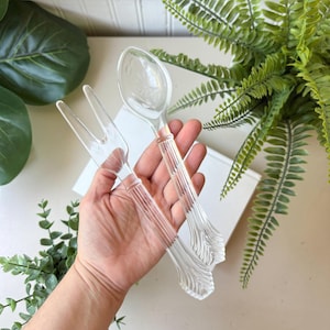 Può includere: Set di cucchiaio e forchetta da portata per insalata in plastica trasparente. Il cucchiaio e la forchetta hanno un design con manico a coste e un'estremità svasata decorativa. Il cucchiaio ha un motivo floreale sulla ciotola. Il set è tenuto in mano.