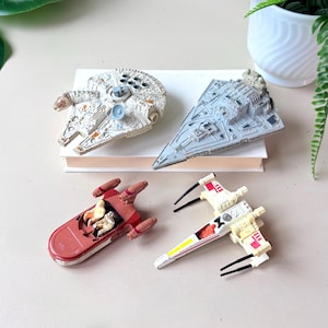 May include: Four Star Wars toy spaceships are arranged on a white surface. The set features a Millennium Falcon, a Star Destroyer, a red speeder, and an X-wing fighter. The toys are detailed with various colours and designs.