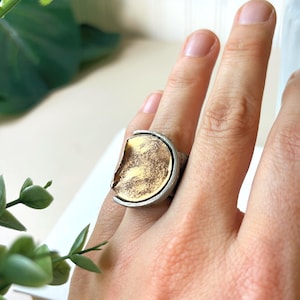 May include: A ring featuring a circular gold-toned centrepiece with a textured, metallic finish. The gold disc is framed by a curved, silver-toned metal band that wraps around the sides, creating a layered, two-tone appearance on the finger.