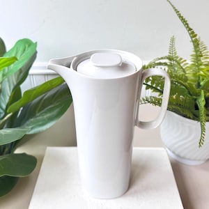 以下が含まれることがあります： 取っ手と蓋が付いた、背の高い白いセラミックピッチャー。ピッチャーは、洗練されたモダンなデザインで、注ぎやすい注ぎ口が付いています。ハンドルは長方形で、蓋は小さく正方形のノブが付いています。飲み物を提供するのに適しています。