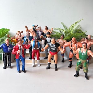 May include: A collection of vintage plastic professional wrestling action figures featuring various characters in colourful ring attire, including one figure in a red blazer and another in a black shirt with the text Magnificent Muraco printed on the chest.