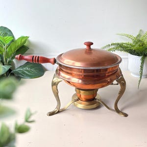 May include: A copper chafing dish with a lid, featuring a long wooden handle with a ribbed texture. The round copper vessel rests on a brass stand with three curved legs and includes a central brass burner component underneath the main bowl.