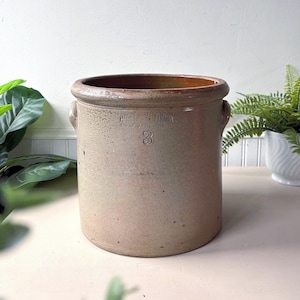 May include: A vintage stoneware crock with a tan exterior and a brown interior. The crock has two small handles and the number "3" is visible. The text "HARRIS LAZAR" and "BAY OF QUINTE, ONT" is also visible.