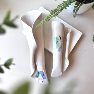 Puede incluir: Dos jarrones de pared de cerámica blanca con un diseño plegado. Un jarrón tiene un diseño floral azul, verde y rosa. El otro jarrón es liso. Los jarrones se exhiben con vegetación.