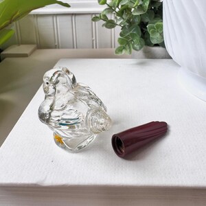 May include: A clear glass bird-shaped perfume bottle with a detached, dark red stopper. The bottle has detailed wings and a small yellow accent. The stopper is a solid, ribbed design.