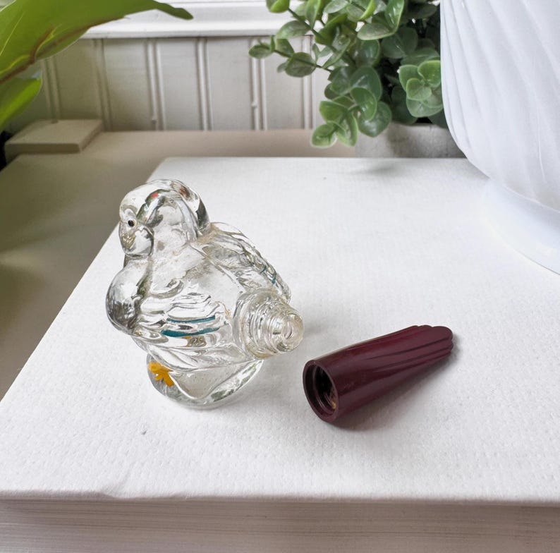 May include: A clear glass bird-shaped perfume bottle with a detached, dark red stopper. The bottle has detailed wings and a small yellow accent. The stopper is a solid, ribbed design.