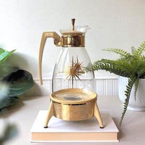 May include: A clear glass carafe with a beige handle and lid, featuring a gold starburst pattern on the body. The carafe sits on a beige and gold metal base with three legs. The lid has a small gold finial, and the base has a matching starburst design.