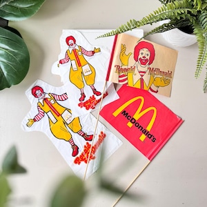 May include: An assortment of vintage McDonald's promotional items. Includes three white kites with Ronald McDonald graphics, a small brown paper sign with Ronald McDonald, and a red flag with the McDonald's logo.