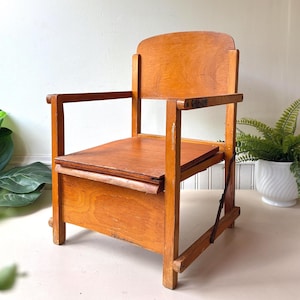 May include: A small wooden chair with a light brown finish, featuring a flat seat, a curved backrest, and two armrests. The chair has a box-like base structure and includes a metal hinge mechanism on the side, showing visible wood grain throughout.