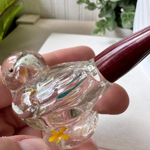 May include: A clear glass bird-shaped perfume bottle with a dark red stopper. The bird has yellow and blue accents and a yellow flower on its base. The bottle is held in a person's hand.