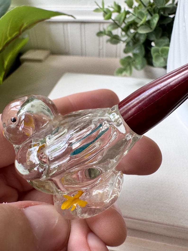 May include: A clear glass bird-shaped perfume bottle with a dark red stopper. The bird has yellow and blue accents and a yellow flower on its base. The bottle is held in a person's hand.