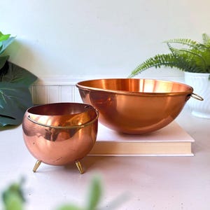 May include: A polished copper bowl with a small side handle and a smaller, spherical copper pot featuring three short legs and a thin metal handle. Both items have a warm, metallic finish and are displayed together as a set of kitchenware.