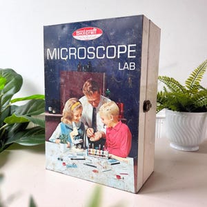 May include: A vintage Skilcraft Microscope Lab box with a blue and white design. The box features an image of a man and two children conducting a science experiment. The text "MICROSCOPE LAB" is prominently displayed on the front.