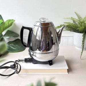 May include: A polished chrome electric percolator coffee pot with a black handle and a clear glass knob on the lid. The body features a vertical embossed logo with the letters SB. It includes a black base and a black power cord with a plug.
