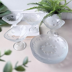 Puede incluir: Un juego de copas y platos de postre de cristal transparente. Las copas tienen forma redondeada con pie y están decoradas con pequeños círculos en relieve. Los platos están apilados y tienen un diseño similar. Plantas verdes en el fondo.