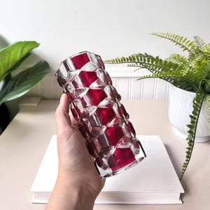 Könnte beinhalten: Eine klare Glasvase mit einem geometrischen, wabenartigen Muster, das rote Akzente in jeder Zelle aufweist. Die Vase ist zylindrisch und wird von einer Hand gehalten, mit einem weißen Buch und Grünzeug im Hintergrund. Die Vase ist etwa 20 cm hoch.