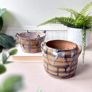May include: Two handmade ceramic pots with handles. The pots are tan with a dark blue plaid pattern. One pot is in the foreground, and the other is in the background. The pots are sitting on a white surface.