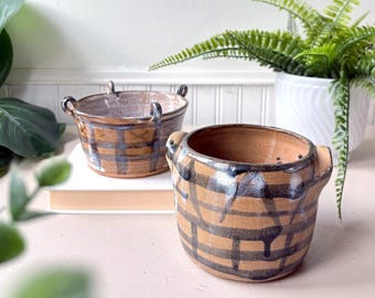 May include: Two handmade ceramic pots with handles. The pots are tan with a dark blue plaid pattern. One pot is in the foreground, and the other is in the background. The pots are sitting on a white surface.