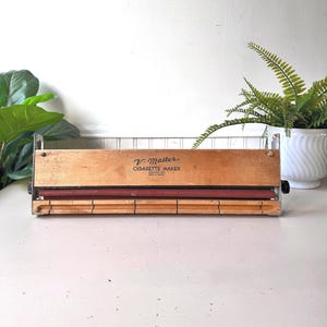 May include: A vintage V-Master cigarette maker, constructed from wood and metal. The wooden body is light brown, featuring a red roller and the text "V-Master CIGARETTE MAKER". It includes a metal wire cage and a black knob on the side.