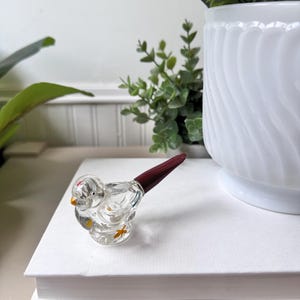 May include: A clear glass bird figurine with a dark red tail and yellow accents. The bird is perched on a stack of white books. A white vase with green plants is in the background.