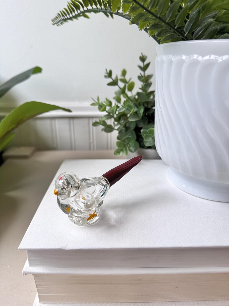 May include: A clear glass bird figurine with a dark red tail and yellow accents. The bird is perched on a stack of white books. A white vase with green plants is in the background.