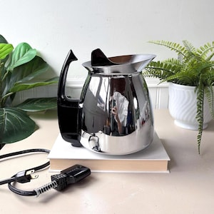 Puede incluir: Una cafetera eléctrica cromada vintage con asa y boquilla negras. La cafetera está sobre una pila de libros. Un cable de alimentación y un enchufe negros están en primer plano. Plantas verdes en el fondo.