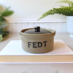 May include: A round ceramic crock with a light tan base and a dark brown lid. The word FEDT is stamped in black capital letters on the front of the container. The vessel features a simple cylindrical shape with a small knob on the center of the lid.