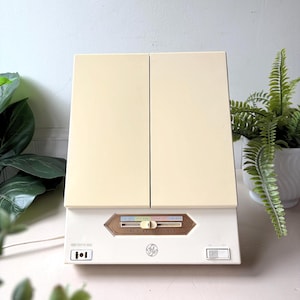 May include: A vintage GE "True Reflections" vanity mirror with a cream-coloured cabinet and two doors. The base is white with a brown and gold accent panel and the GE logo. The mirror has a light and a switch. Decorative plants are in the background.