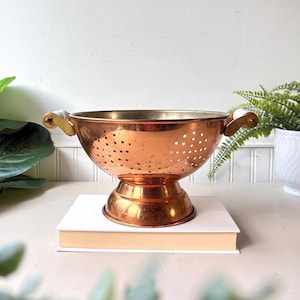 May include: A polished copper colander featuring a wide, circular bowl with numerous drainage holes, a sturdy pedestal base, and two side handles with white accents. The metallic surface reflects light, highlighting the warm, reddish-brown tone.