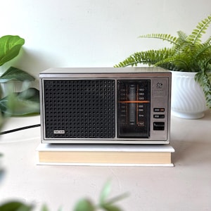 Puede incluir: Una radio FM/AM General Electric rectangular con panel frontal metálico plateado, rejilla de altavoz negra y pantalla de sintonización de frecuencia vertical. El dispositivo cuenta con botones selectores AM y FM y un cable negro.