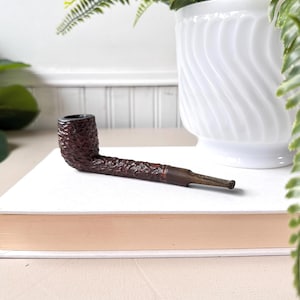 Peut inclure: Une pipe à tabac marron avec un foyer texturé et une longue tige fine. La pipe est posée sur une surface blanche, avec un pot décoratif blanc et de la verdure en arrière-plan. Un accessoire de fumeur classique.