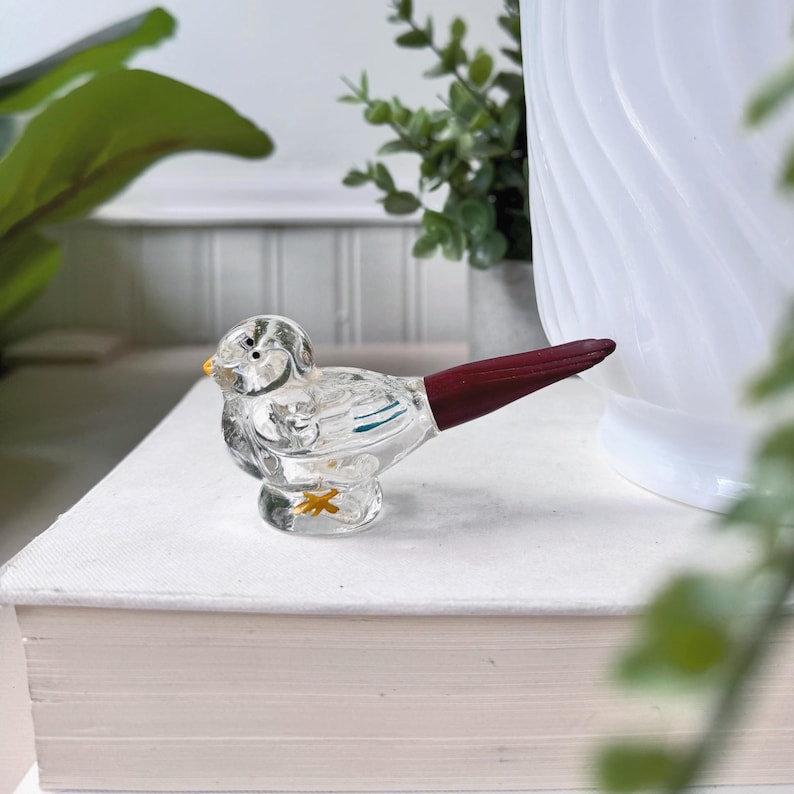 May include: A clear glass bird figurine with a dark red tail and yellow beak. The bird has etched details and sits on a small base. The figurine is displayed on a white surface.