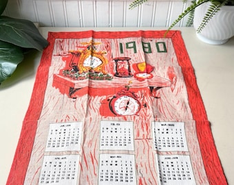 May include: A vintage calendar towel from 1980 with a red border and a wood-grain design. The top features a clock, hourglass, and other decorative elements. Monthly calendars are displayed below, with the months of the year printed in black.